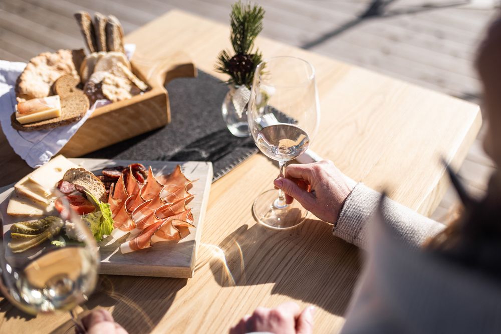 Tagliere con affettati, formaggi, pane e due persone che tengono calici di vino bianco su un tavolo di legno all'aperto