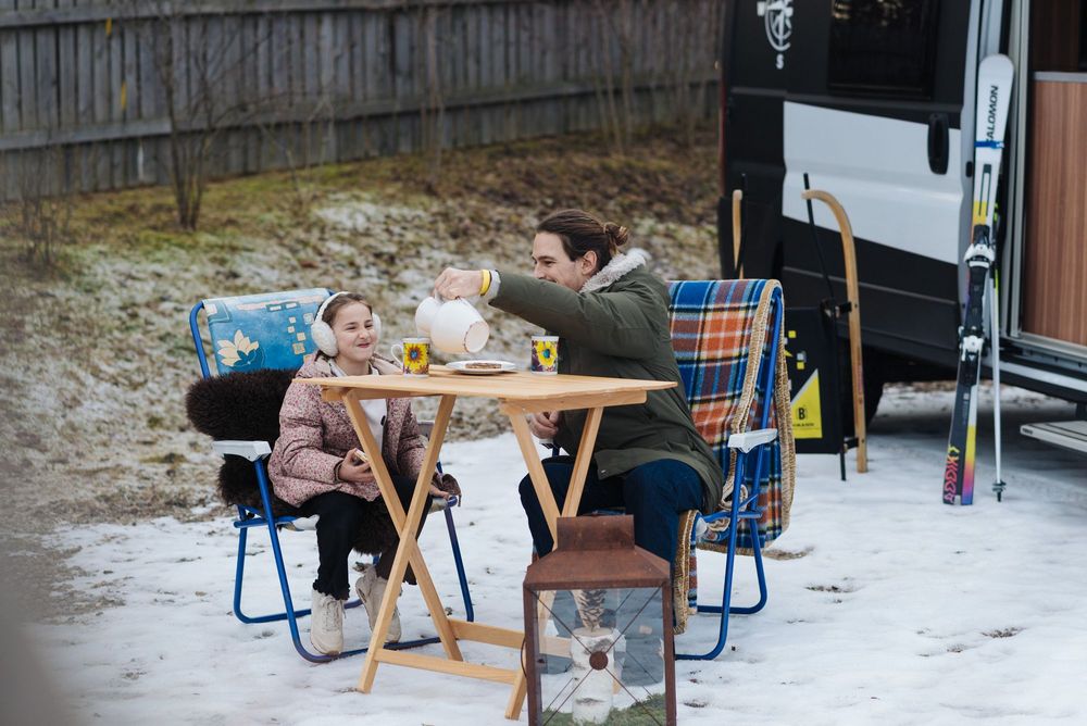 Padre e figlia seduti all'aperto sulla neve con bevande calde e biscotti vicino a un camper con sci