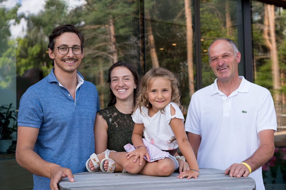 A smiling family of four posing outdoors, with a young girl sitting on a wooden table.