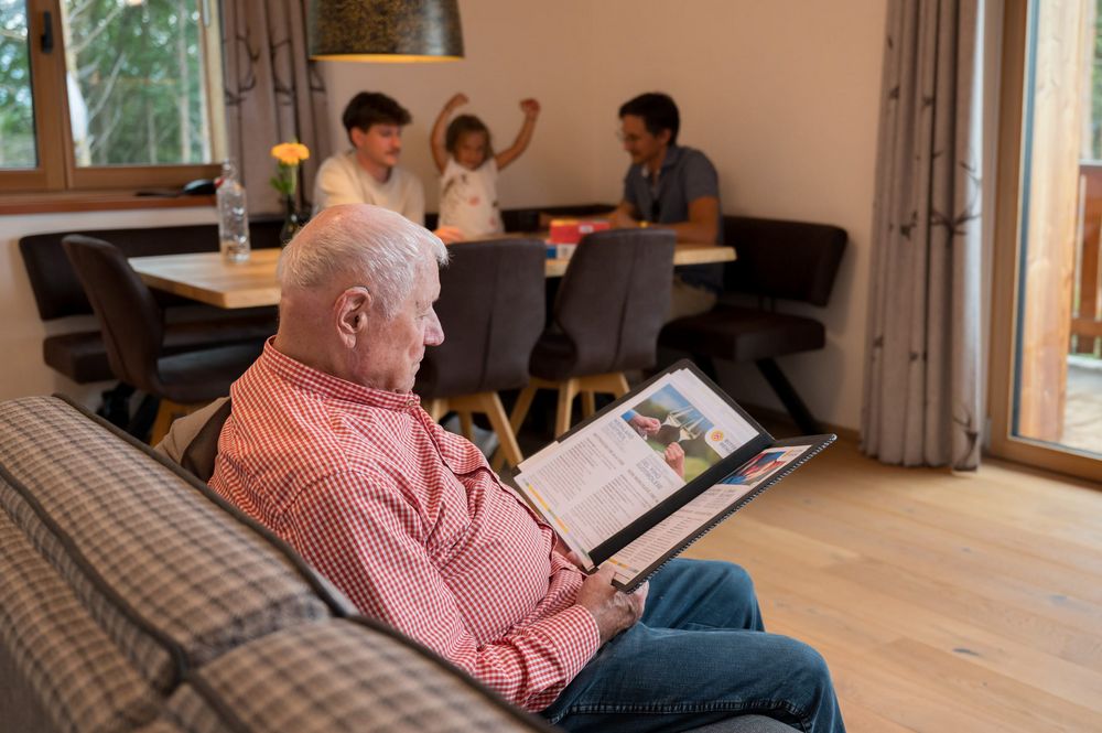 Älterer Mann liest in einem Ordner, während im Hintergrund zwei Männer und ein Kind am Tisch sitzen.