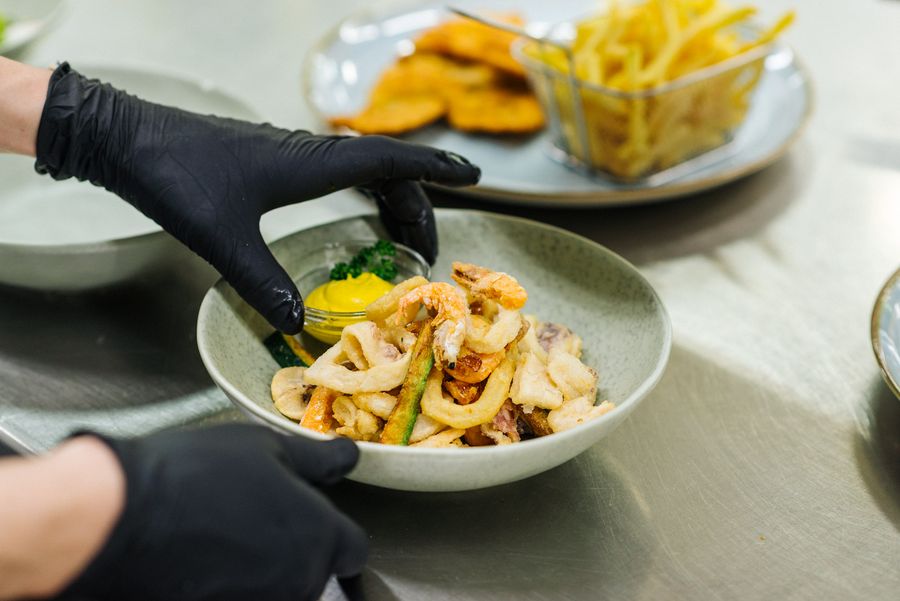 Person mit schwarzen Handschuhen serviert frittierte Meeresfrüchte mit Sauce in einer Schüssel.