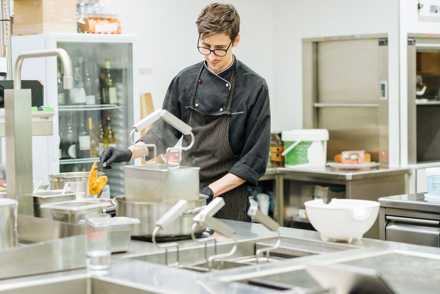 Koch in schwarzer Uniform und Schürze frittiert Lebensmittel in einer professionellen Küche.