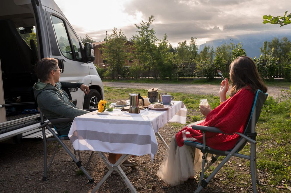 Due persone sedute all'aperto, vicino a un camper, che pranzano con una vista naturale