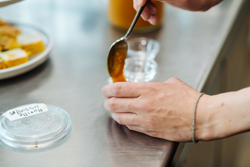 Hand hält Löffel mit orangefarbener Sauce über einem kleinen Glasbehälter auf einer Metalloberfläche.