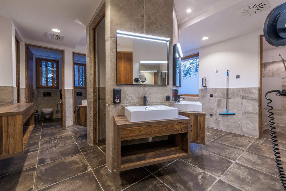 Bagno moderno con pavimento in piastrelle scure, lavandini bianchi su mobili in legno e specchi illuminati.
