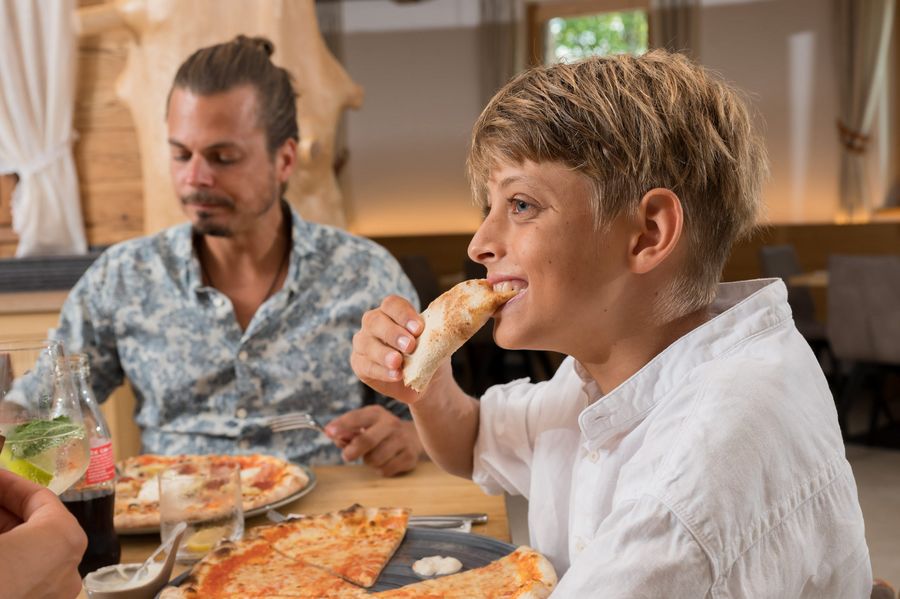 Junger Mensch isst Pizza, im Hintergrund sitzt ein Mann mit Hemd und isst ebenfalls Pizza.