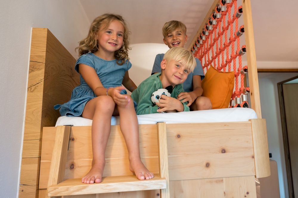 Drei lachende Kinder sitzen auf einem Hochbett aus Holz, eines hält ein Plüschtier.