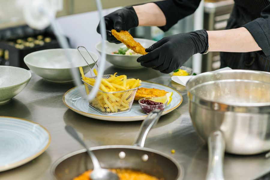 Person in schwarzer Kleidung legt paniertes Fischfilet auf Teller mit Pommes frites und Soße in einer Küche