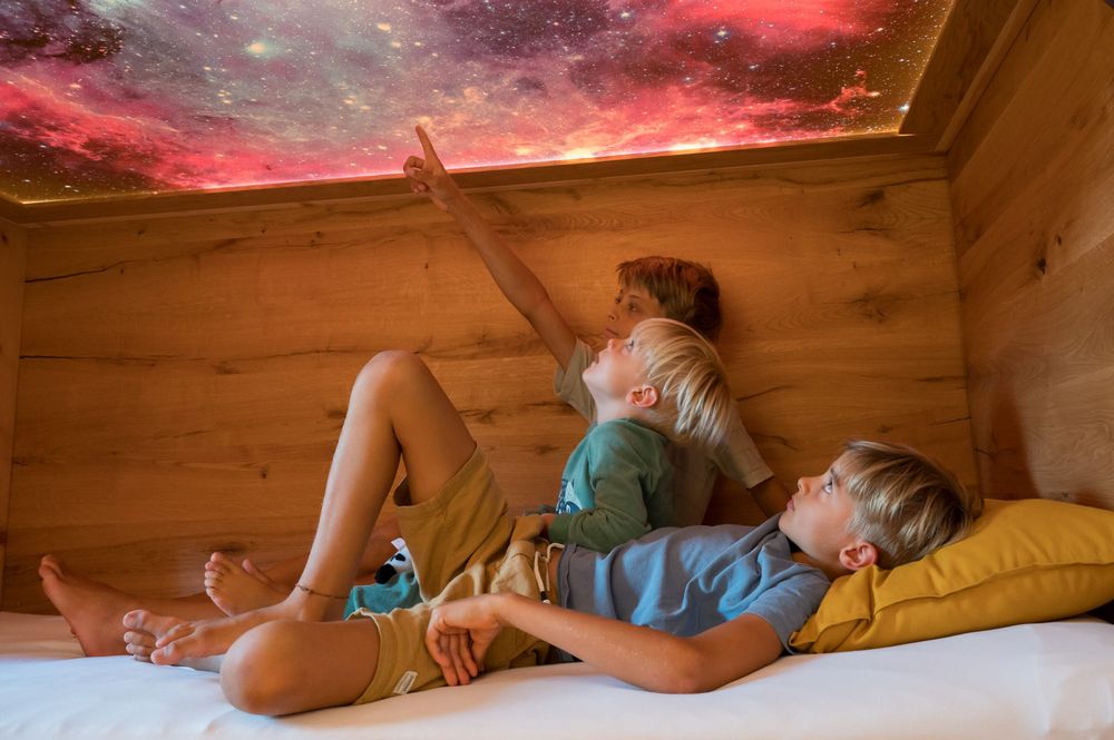 Drei Kinder liegen auf einem Bett und schauen an eine Decke mit einem Sternenhimmel-Motiv.