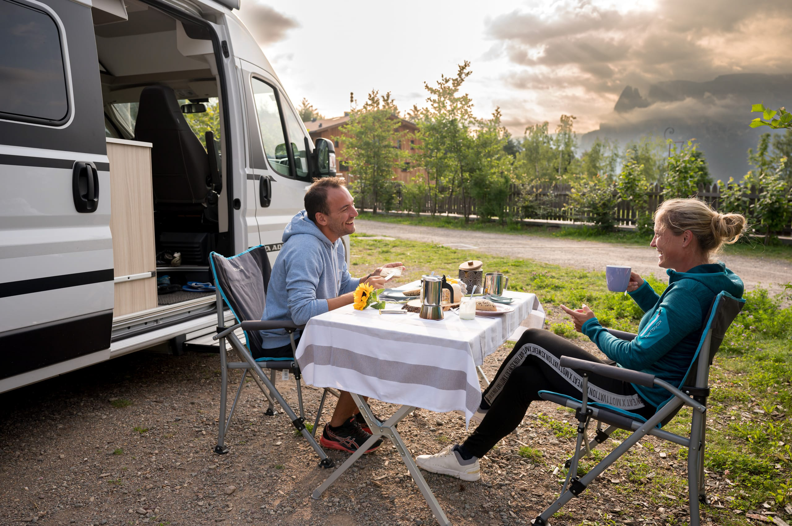 Zwei Personen sitzen an Campingtisch neben Wohnmobil und genießen Kaffee im Freien.