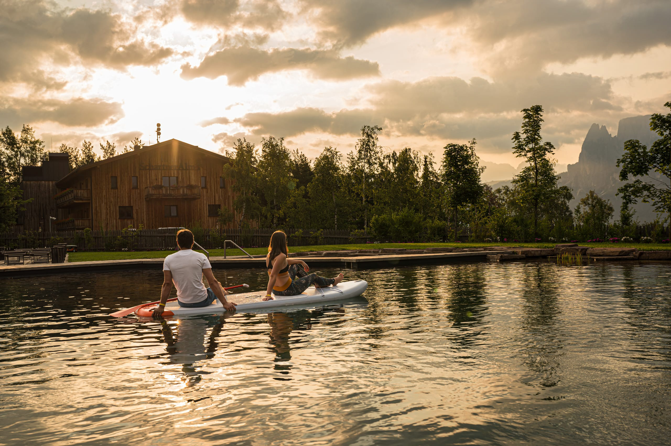 Zwei Personen sitzen auf einem Stand-Up-Paddle-Board auf ruhigem Wasser bei Sonnenuntergang vor einem Holzhaus.